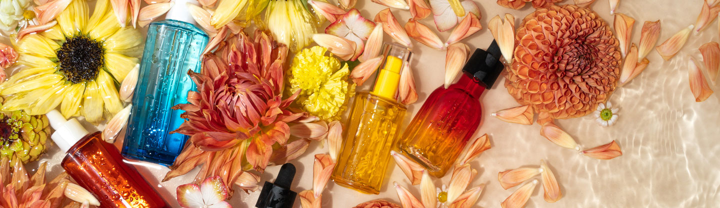 Cosmetic bottles and flowers floating in water. Face serums with sunflowers, dahlias and carnations on beige background.